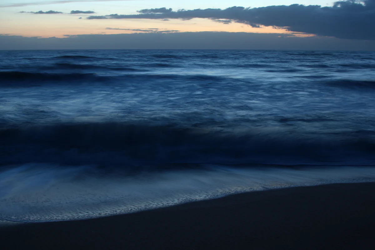 Waves blurred into soft blue motion on a dark beach at dusk, under a band of peach light and cloud