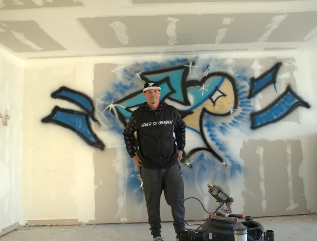 Vanilla Ice in a Never Surrender hoodie, hands on his hips, in front of his spray painted tag inside the beach house