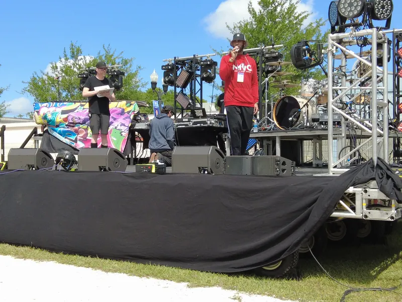 An outdoor stage in daylight with lighting rigs and crew setting up