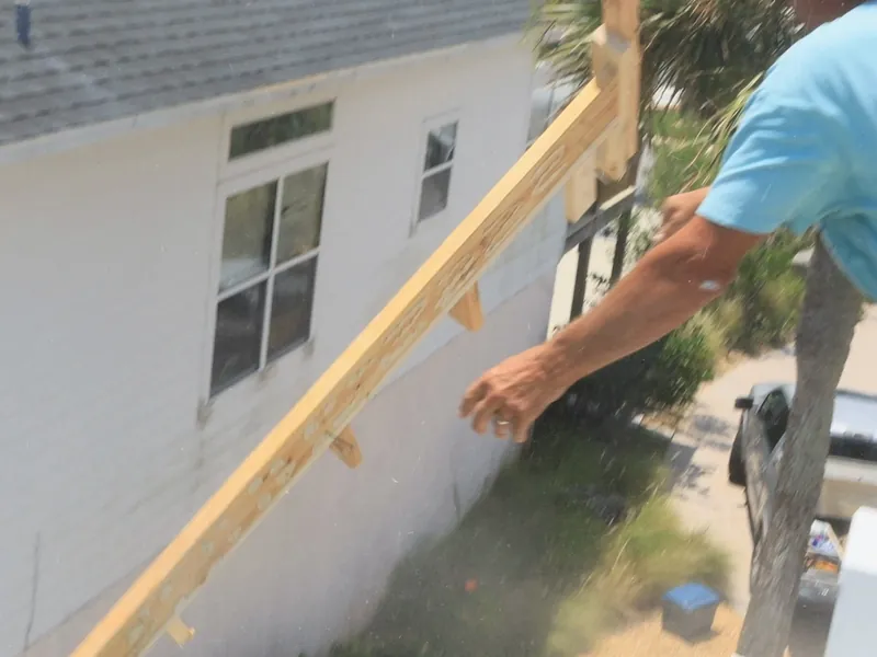 A worker on an upper deck passing a length of lumber down beside a white house with a shingle roof