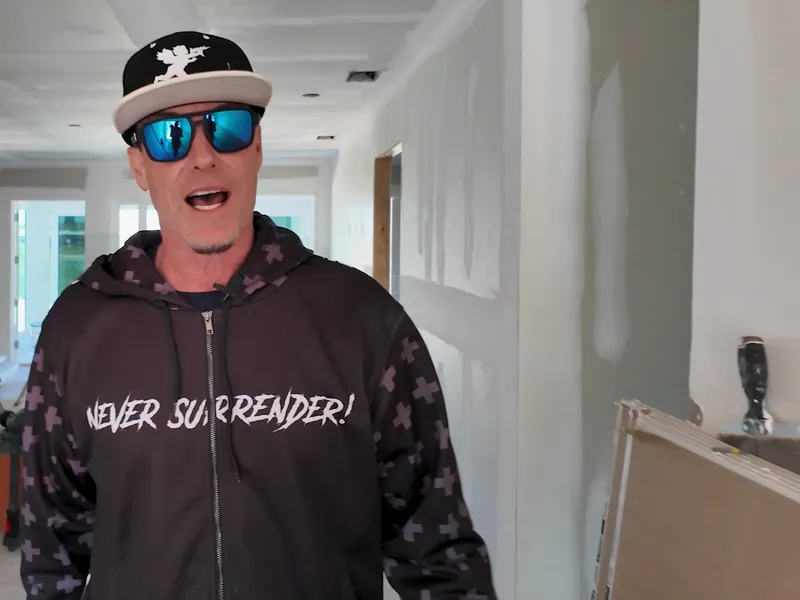 Vanilla Ice talking to the camera inside the house, with bare drywall and building supplies behind him